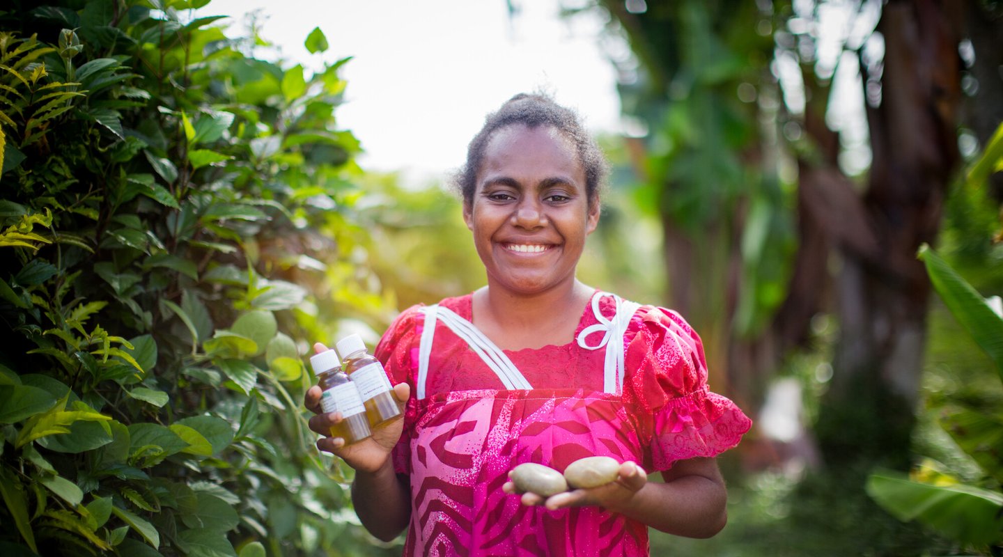 Nelsie, a smiling woman in a pink top, holds up the soaps and oils that she has made as part of her small business in Vanuatu.