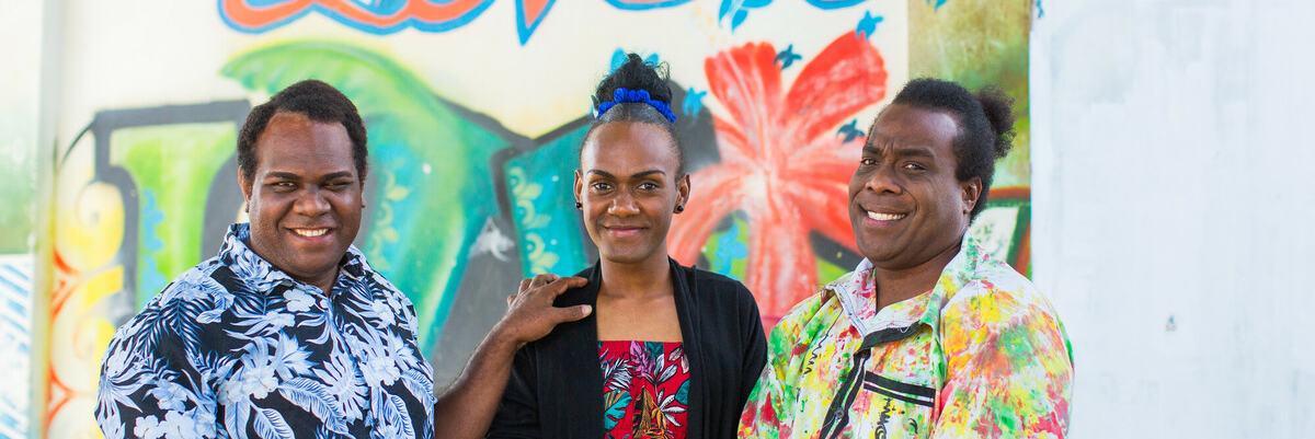 Three people are stood outside the VPride office in front of a colourful wall with the word Love painted on it.