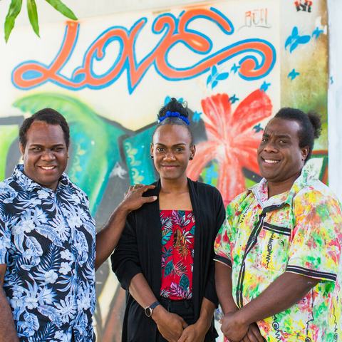 Three people are stood outside the VPride office in front of a colourful wall with the word Love painted on it.