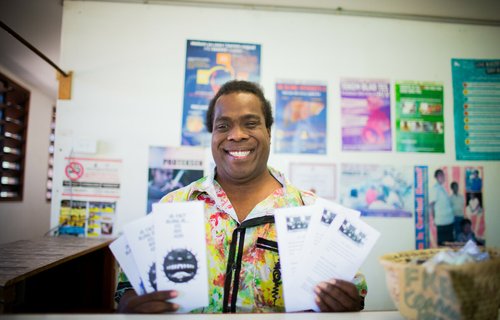 A smiling man from Vanuatu Pride holds up sexual health pamphlets at an outreach centre, with educational posters visible on the wall behind him.