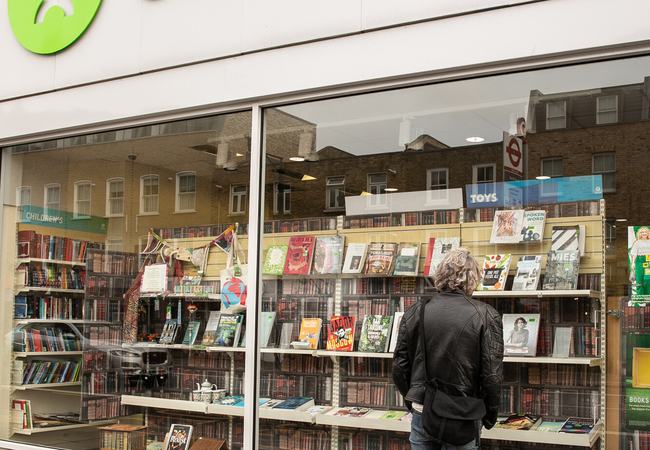 An exterior shot of a man standing looking into the window of an Oxfam shop , featuring a large selection of donated books in the window display.