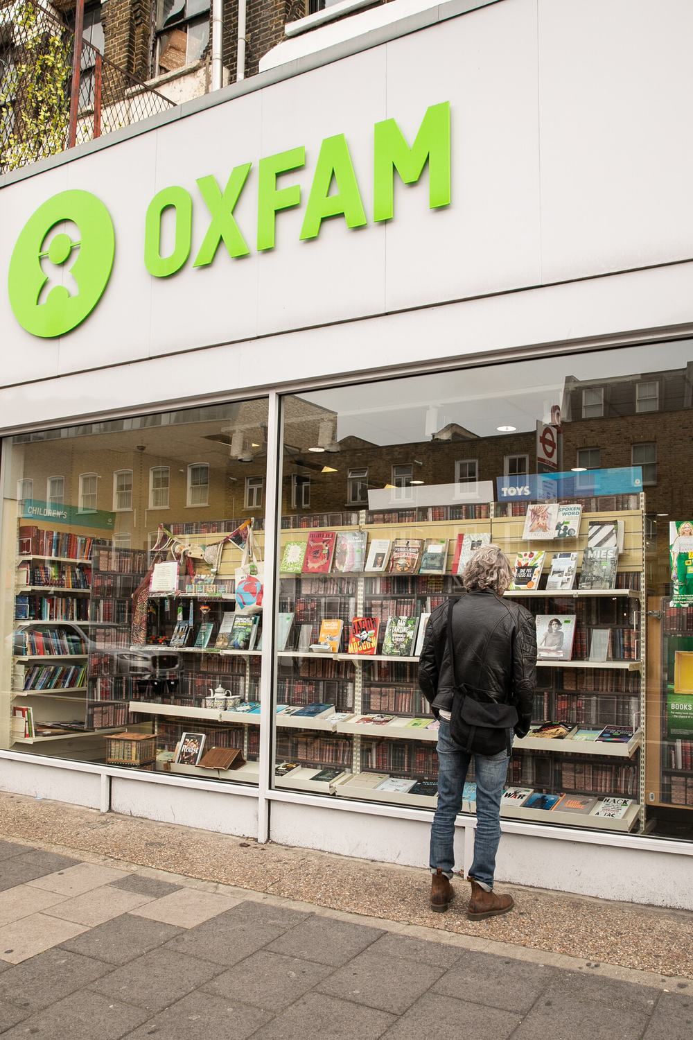 An exterior shot of a man standing looking into the window of an Oxfam shop , featuring a large selection of donated books in the window display.