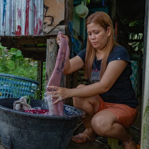 A young woman is crouched outside her home washing some clothes in a large tub.