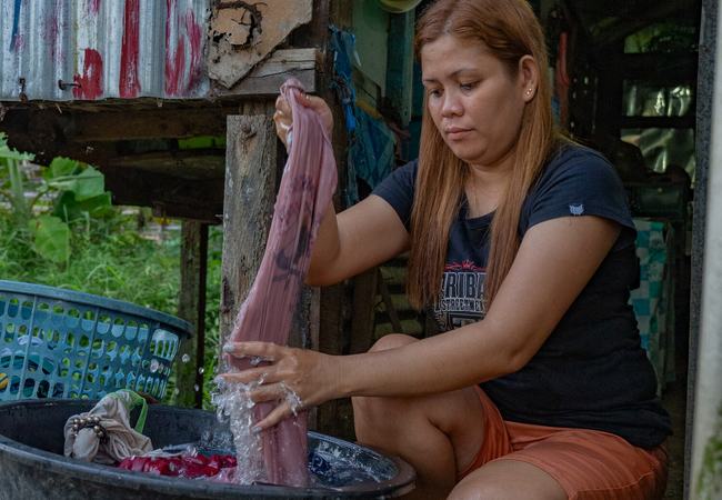 A young woman is crouched outside her home washing some clothes in a large tub.