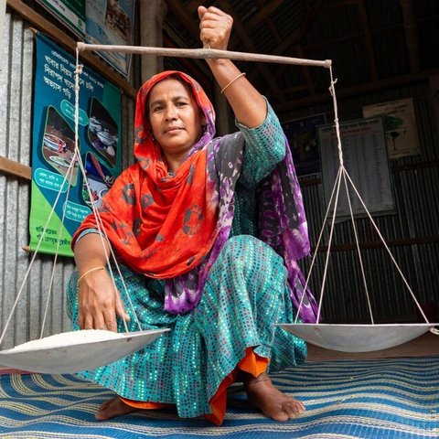 A woman holding up a pair of old-fashioned scales, balancing a bowl on either side. One of the two bowls is full of rice.