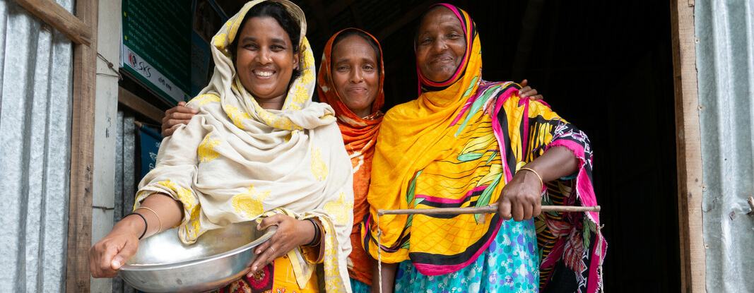 Three women standing in the doorway of their local foodbank in South Gabindi, Bangladesh. The woman on the left is holding a large metal bowl and the woman on the right is holding a large set of balance scales.