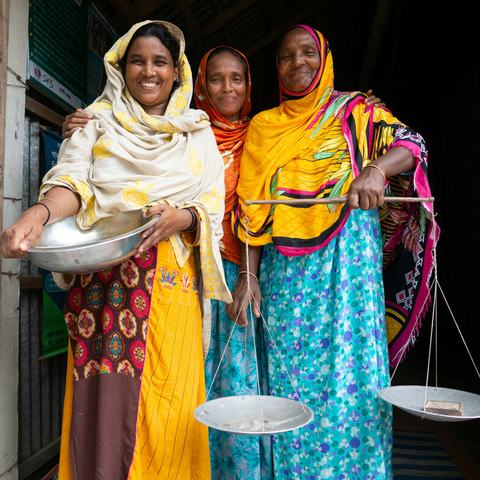Three women standing in the doorway of their local foodbank in South Gabindi, Bangladesh. The woman on the left is holding a large metal bowl and the woman on the right is holding a large set of balance scales.