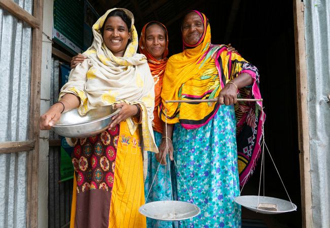 Three women standing in the doorway of their local foodbank in South Gabindi, Bangladesh. The woman on the left is holding a large metal bowl and the woman on the right is holding a large set of balance scales.