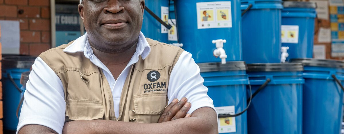 A man in a white cap and a tan-coloured Oxfam utility vest stands in front of a stack of blue water containers in Lusaka, Zambia.