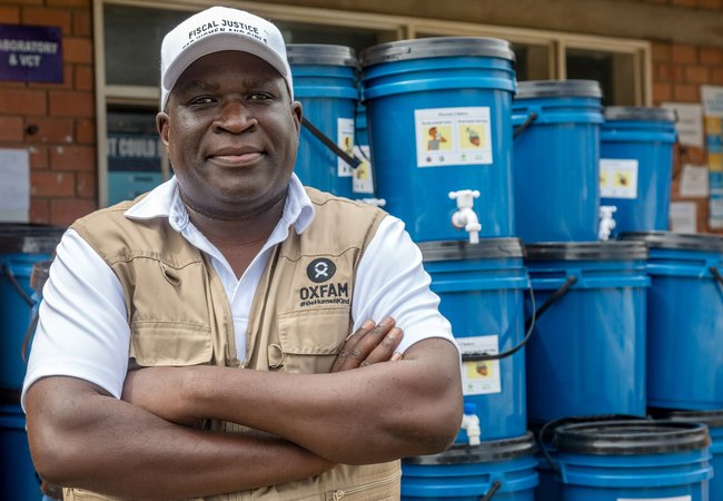 A man in a white cap and a tan-coloured Oxfam utility vest stands in front of a stack of blue water containers in Lusaka, Zambia.
