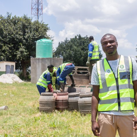 A Zambian man in a hi vis Oxfam jacket with men at work in the background.