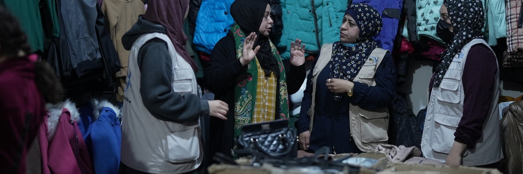 A group of women in hijabs stand talking to each other in a basic shop with childrens clothes hanging from hangers on the walls around them.