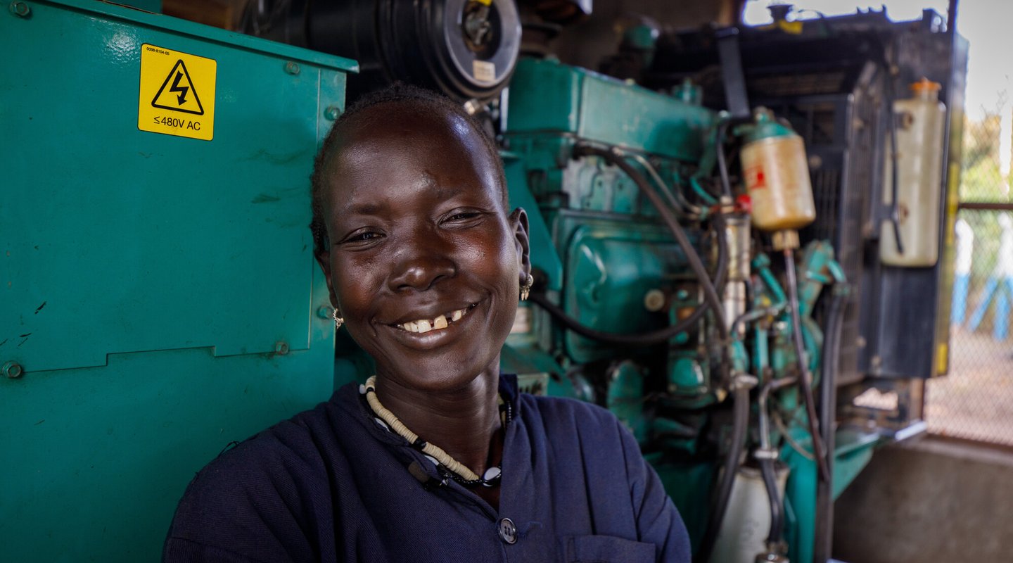 Ethiopian woman Nyathak stands smiling in a blue boilersuit next to green machinery