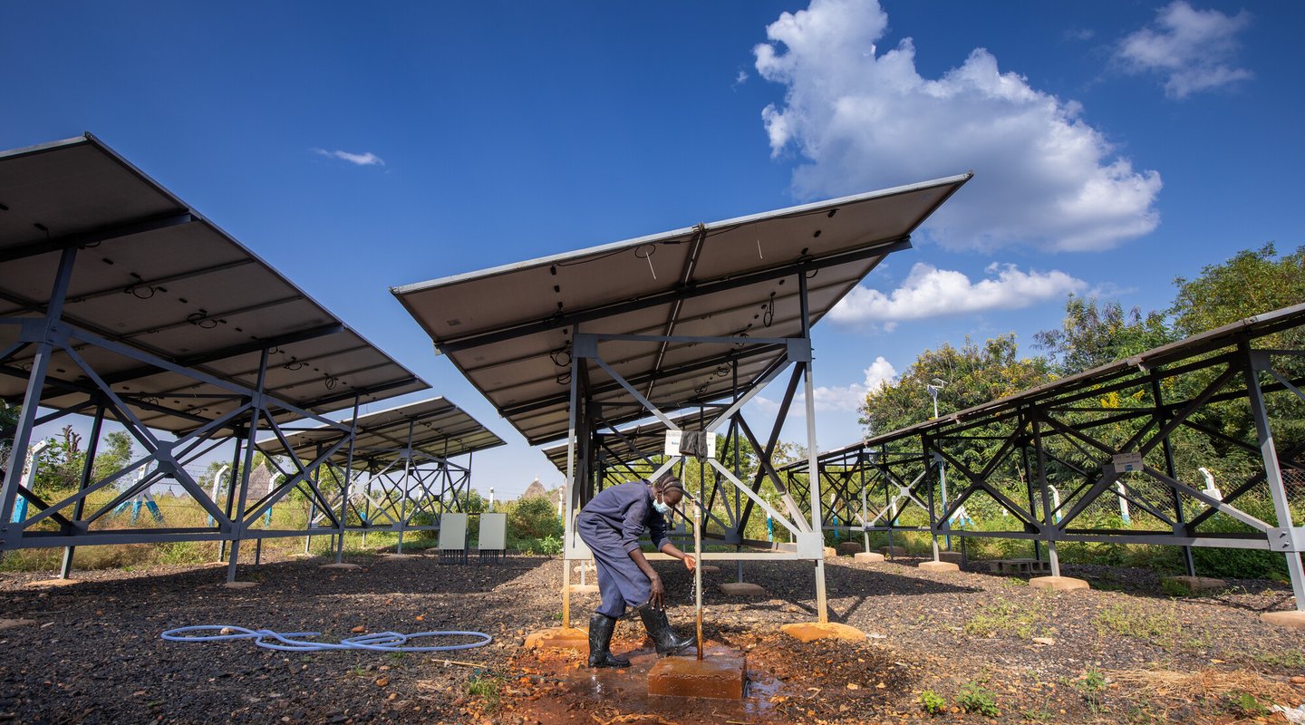 An Ethiopian woman wearing a dress surgical mask beds down to fix something next to massive solar panels that tower behind her.