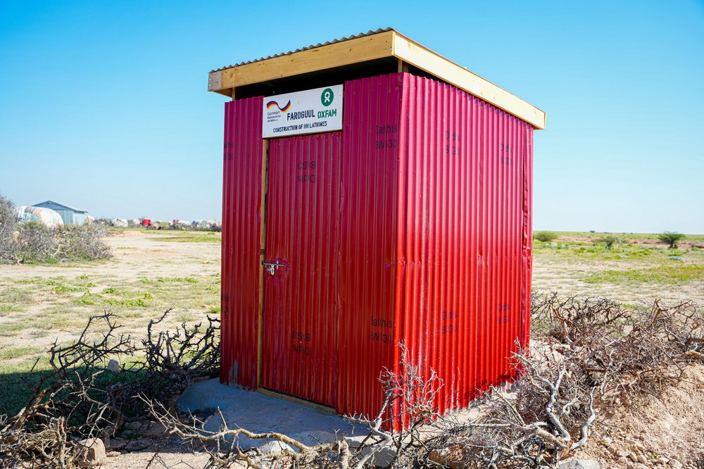 A red latrine outhouse stands in a field with other buildings in the distance.