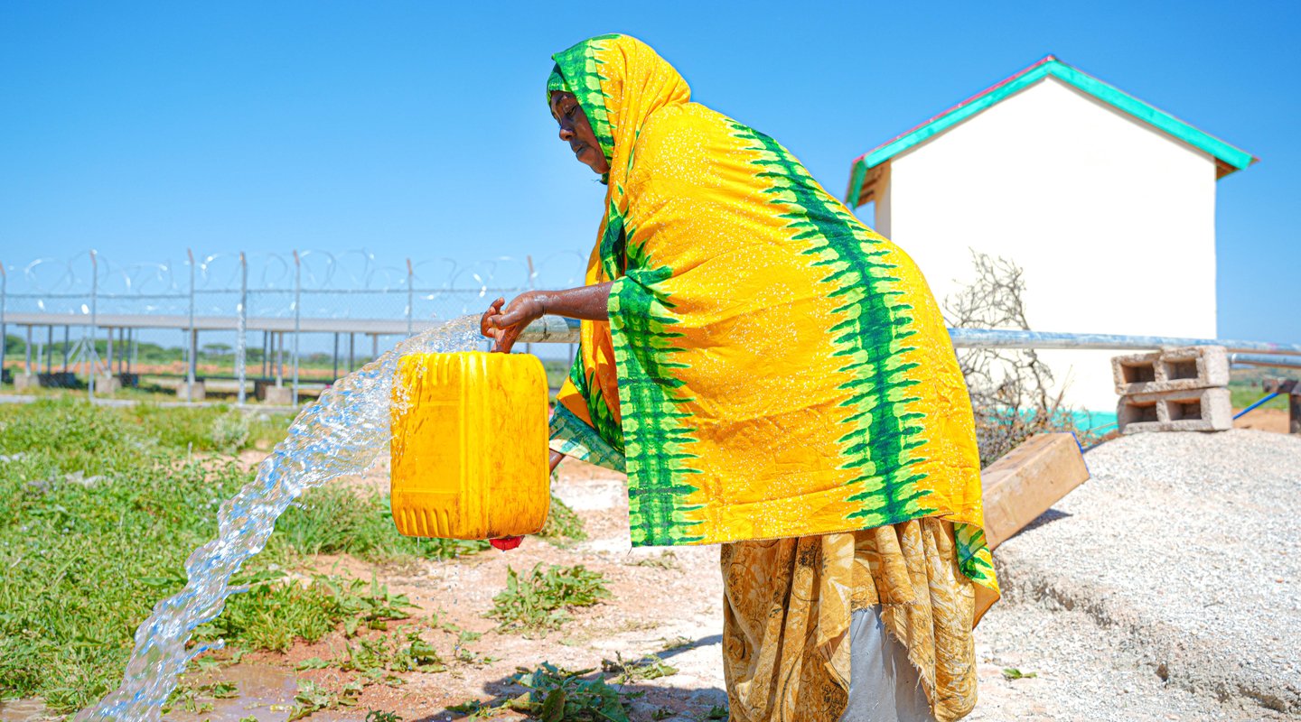 A woman in Somaliland wearing yellow with green stripes pours water from a streaming tap into a jerry can.