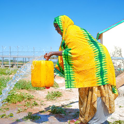 A woman in Somaliland wearing yellow with green stripes pours water from a streaming tap into a jerry can.