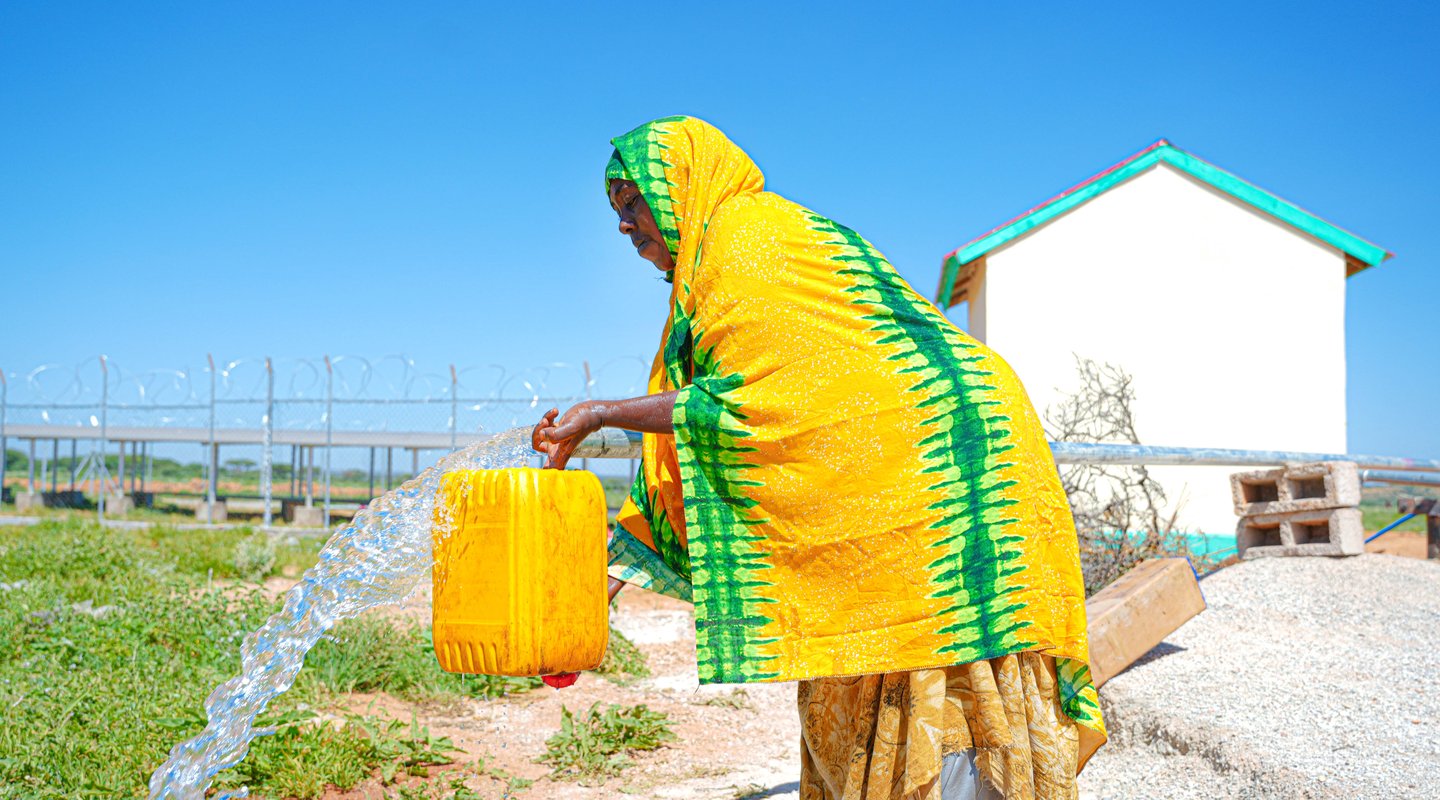 A woman in Somaliland wearing yellow with green stripes pours water from a streaming tap into a jerry can.