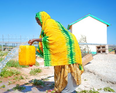 A woman in Somaliland wearing yellow with green stripes pours water from a streaming tap into a jerry can.