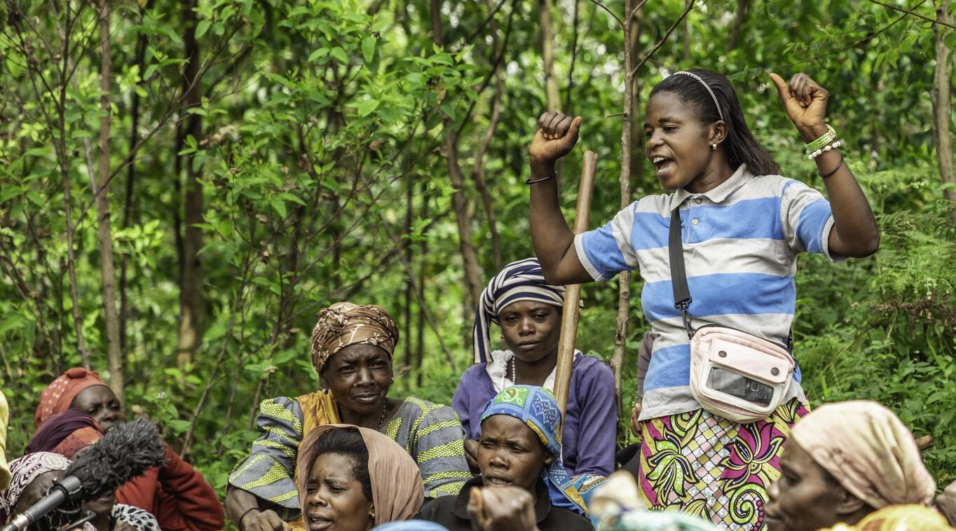 a woman stands amongst other women, sitting.