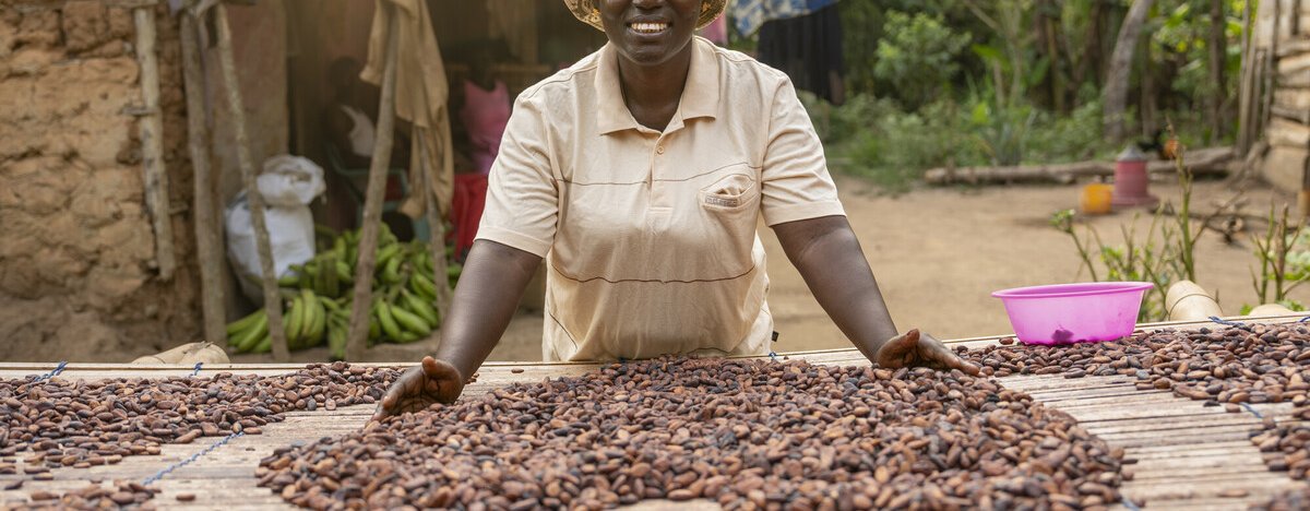 A woman smiles while spreading cocoa beans to dry on a raised platform outside her home. The scene is a rural village with produce, laundry, and greenery in the background.