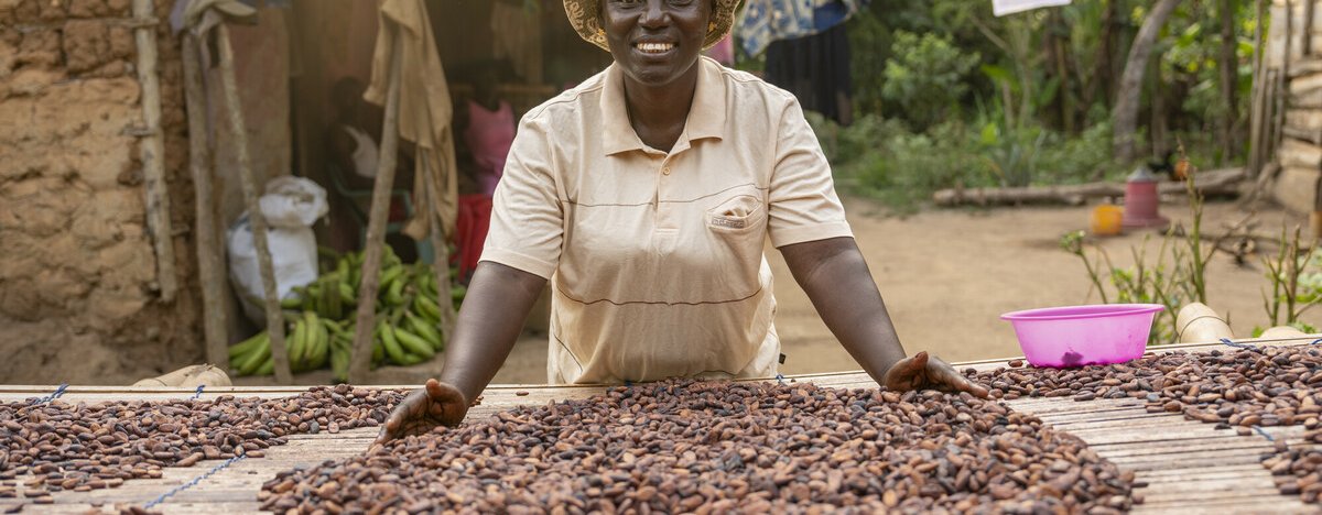A woman smiles while spreading cocoa beans to dry on a raised platform outside her home. The scene is a rural village with produce, laundry, and greenery in the background.
