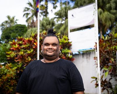 A person standing in front of a natural backdrop, an LGBTQIA+ flag can be seen on a flagpole in the background.