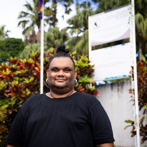 A person standing in front of a natural backdrop, an LGBTQIA+ flag can be seen on a flagpole in the background.