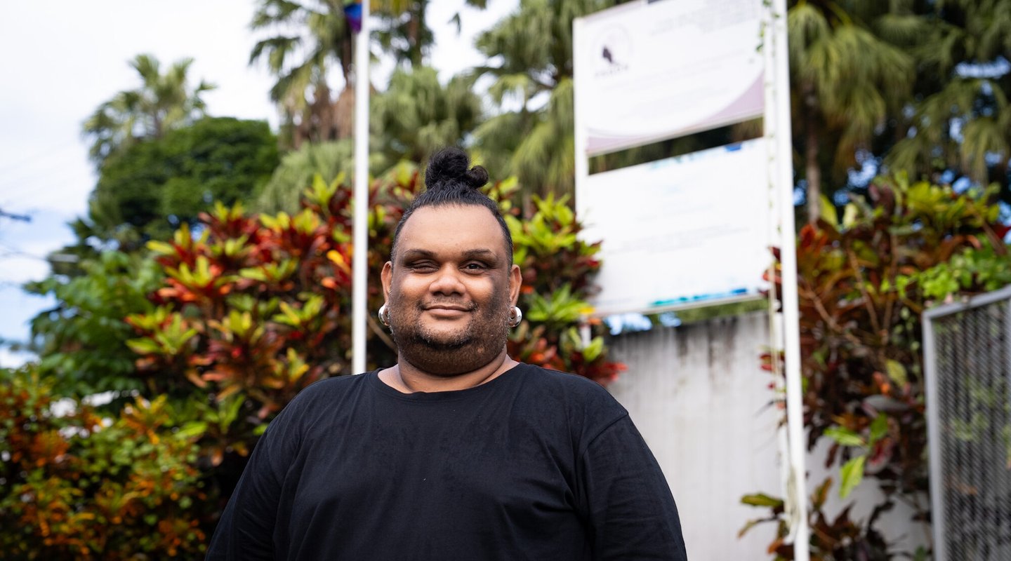 A person standing in front of a natural backdrop, an LGBTQIA+ flag can be seen on a flagpole in the background.