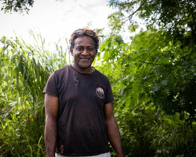 Poasa (he/him), also known as Porsha, stands in front of lush greenery at the PRIDE communal farm in Nakavu village, Nadi, Fiji, part of a project supporting LGBTQIA+ “Queerpreneurs” with agricultural and financial training.