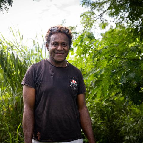 Poasa (he/him), also known as Porsha, stands in front of lush greenery at the PRIDE communal farm in Nakavu village, Nadi, Fiji, part of a project supporting LGBTQIA+ “Queerpreneurs” with agricultural and financial training.
