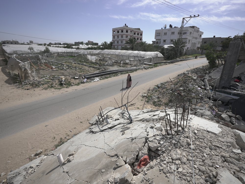 A woman wearing a hijab and abaya walks alone along a road with bombed buildings either side of it.