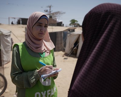 A woman with a pink hijab and green Oxfam vest holds a pen and paper and stands talking to a woman in black hijab by some tents on dusty ground.