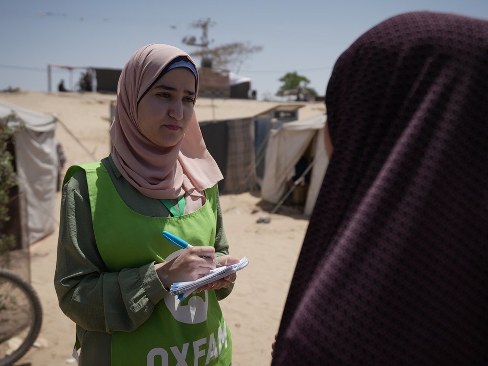 A woman with a pink hijab and green Oxfam vest holds a pen and paper and stands talking to a woman in black hijab by some tents on dusty ground.