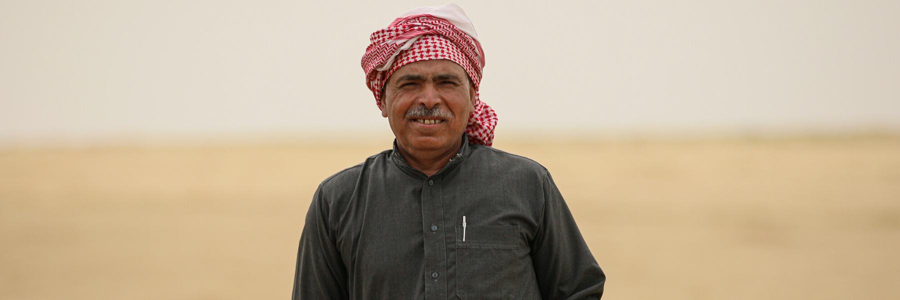 A Syrian man in a red and white keffiyeh stands in a field holding a handful of crops.