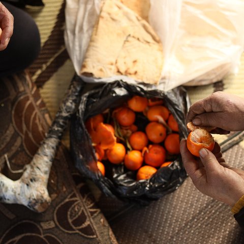 A shot from above of two people peeling madarin oranges above a basket and bread.