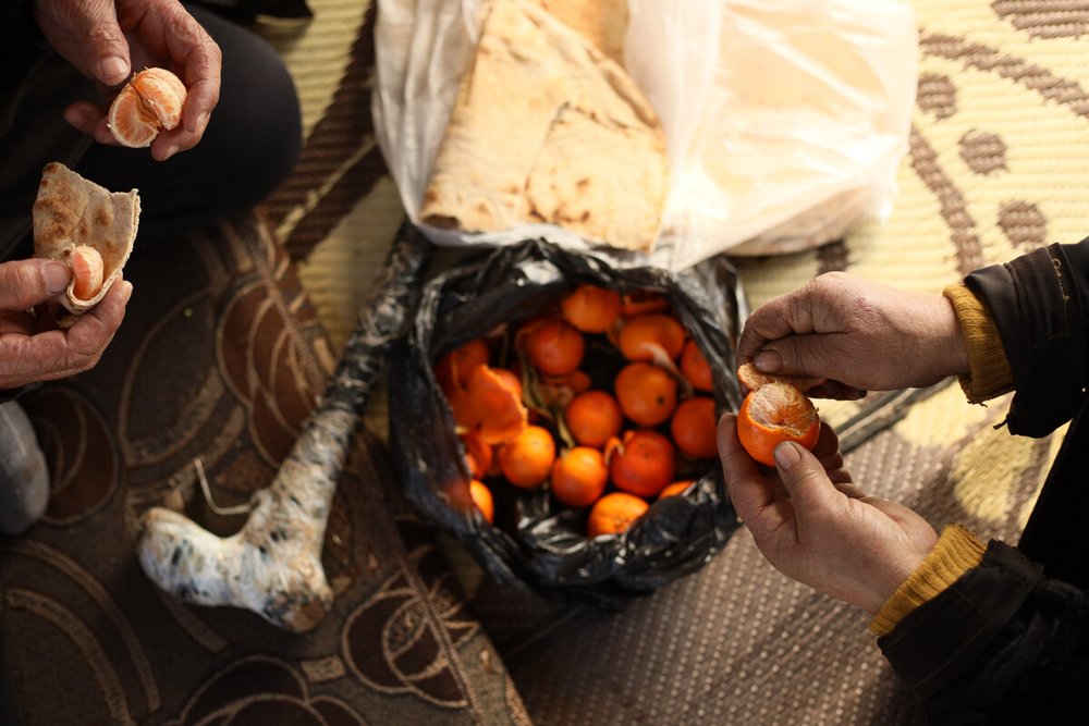 A shot from above of two people peeling madarin oranges above a basket and bread.