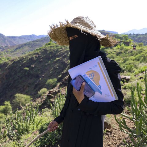 Person wearing a wide straw hat and dark clothing standing outdoors on a green hillside, holding a smartphone and documents with a printed logo. The background shows lush vegetation, cacti, and distant mountains under a clear sky.