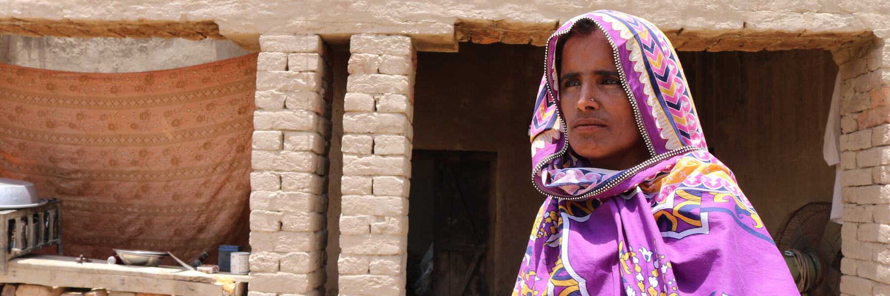 A woman in a purple and yellow head scarf stands outside her home in Pakistan.
