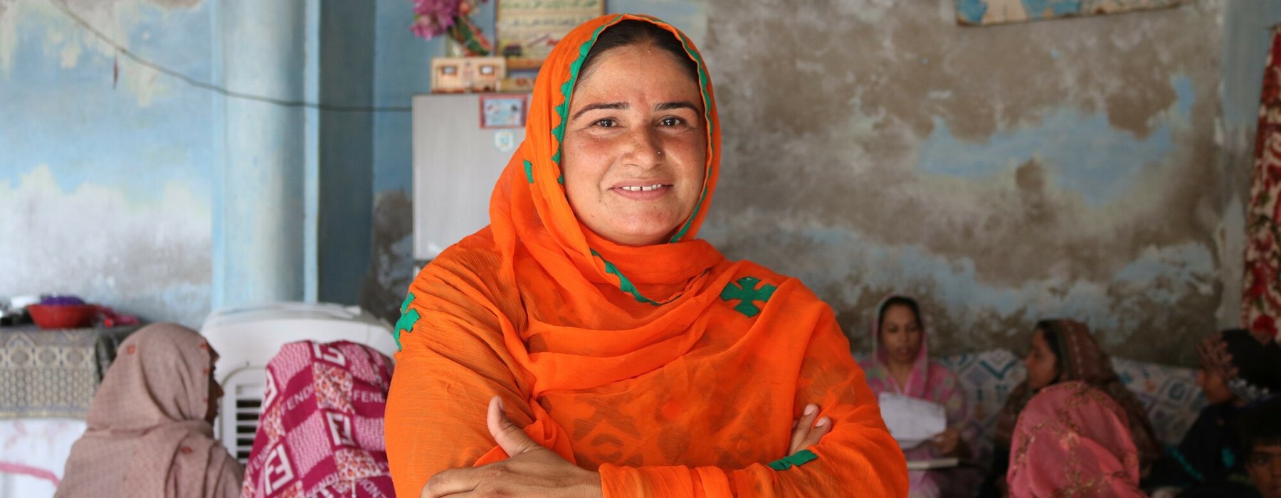 Nazia, a woman from Pakistan wearing an orange scarf, smiles and crosses her arms.
