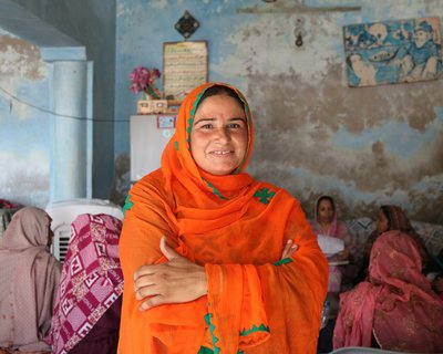 Nazia, a woman from Pakistan wearing an orange scarf, smiles and crosses her arms.