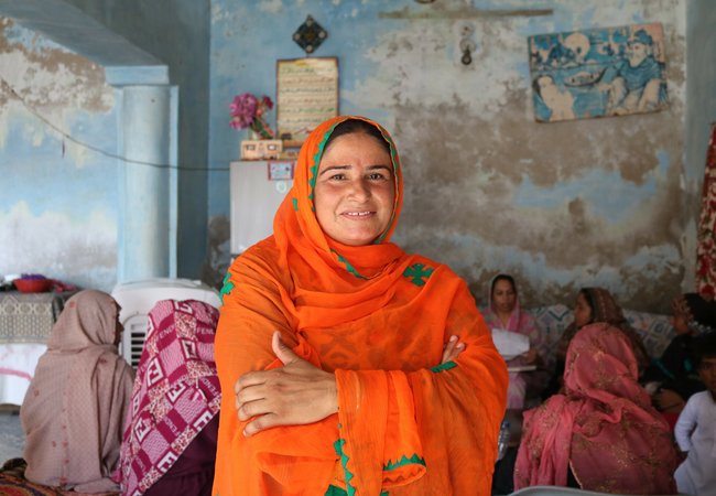 Nazia, a woman from Pakistan wearing an orange scarf, smiles and crosses her arms.