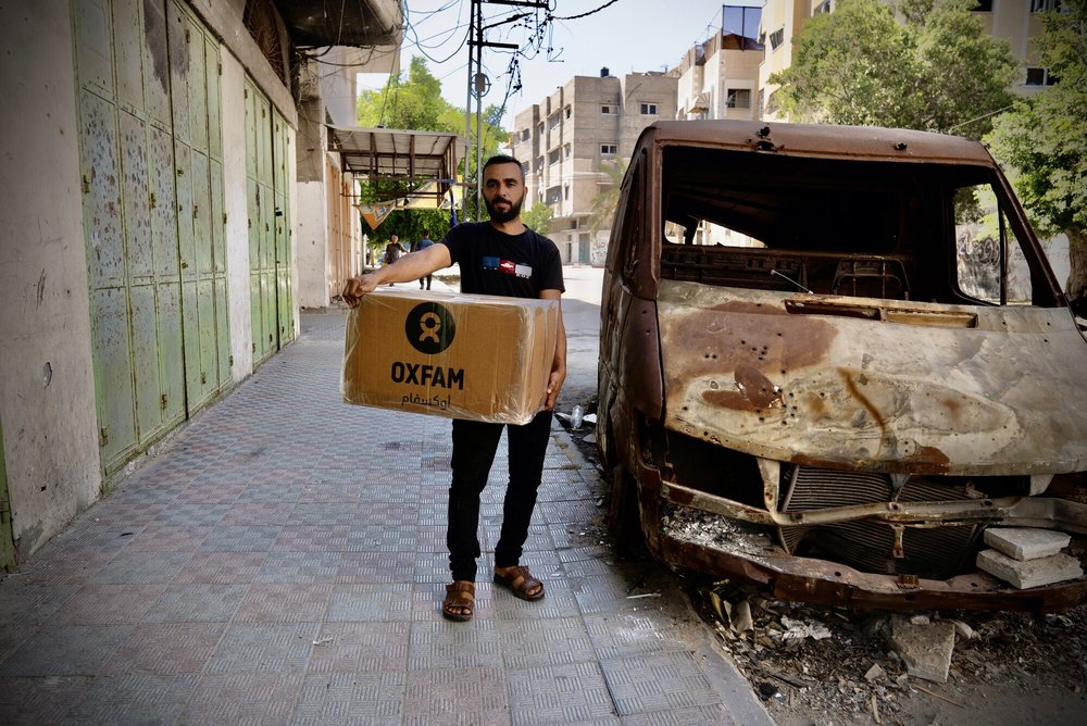 Raed Mansour is holding the Oxfam food parcel distributed among displaced people in Gaza. He stands next to a burnt out car.