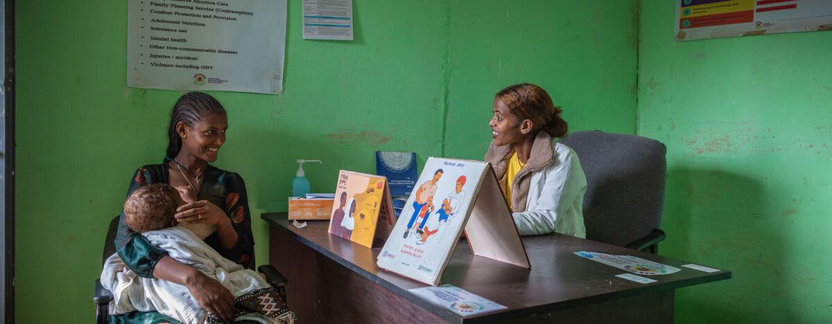 Atsede Bere is sat cradling her young child and smiling at Nurse Bethlehem Feleke who is sat on the opposite side of a desk. The clinic is bright green with posters on the wall and there are promotional healthcare leaflets on the desk.