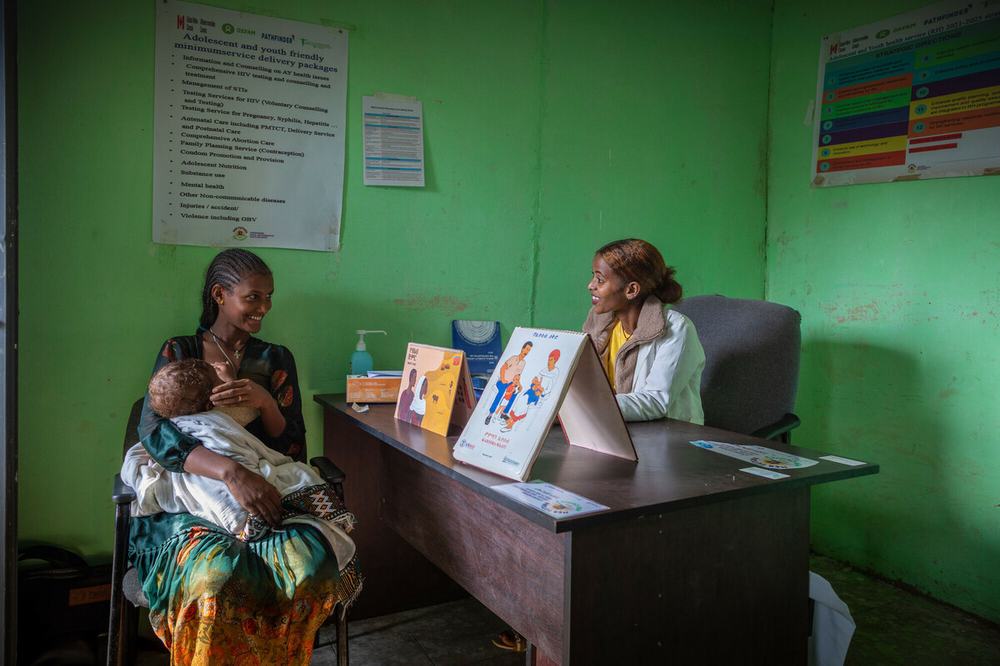 Atsede Bere is sat cradling her young child and smiling at Nurse Bethlehem Feleke who is sat on the opposite side of a desk. The clinic is bright green with posters on the wall and there are promotional healthcare leaflets on the desk.