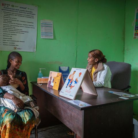 Atsede Bere is sat cradling her young child and smiling at Nurse Bethlehem Feleke who is sat on the opposite side of a desk. The clinic is bright green with posters on the wall and there are promotional healthcare leaflets on the desk.