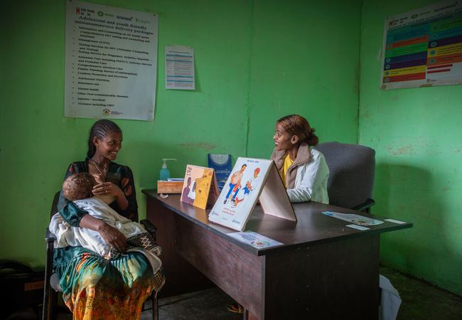 Atsede Bere is sat cradling her young child and smiling at Nurse Bethlehem Feleke who is sat on the opposite side of a desk. The clinic is bright green with posters on the wall and there are promotional healthcare leaflets on the desk.
