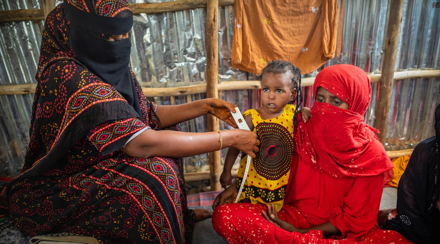 A woman wearing a red and yellow head scarf looks past the camera. Behind her are three other women sitting along the same wall as her.