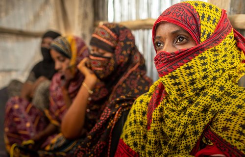 A woman wearing a yellow and red head scarf looks past the camera into the distance. Next to her in the background are three other women. They are all sitting against a wall.