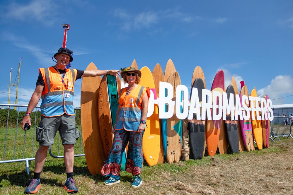 Two Oxfam Festival Stewards standing by a Boardmasters sign post made from surf boards